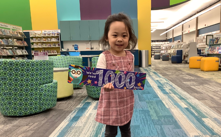 Toddler in pink plaid dress holding up 1,000 Books Before Kindergarten completion sign