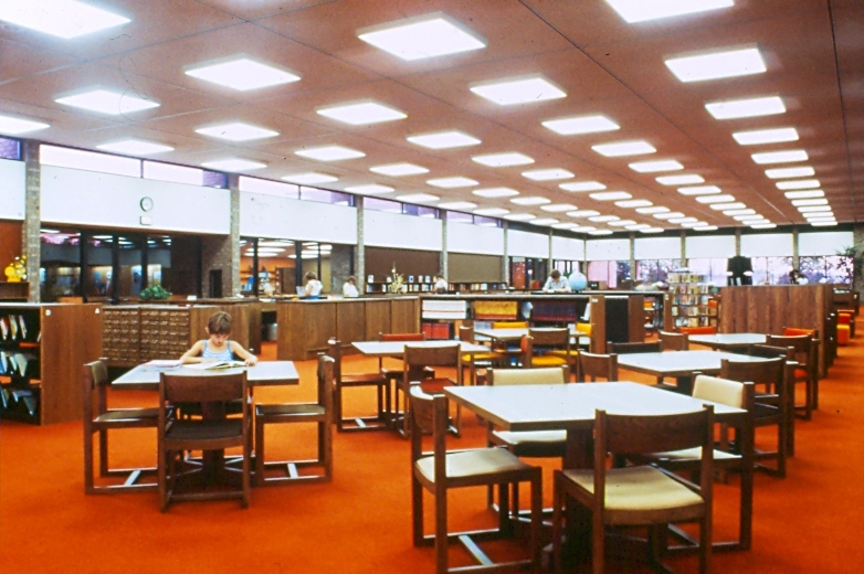 1970s library interior with study desks on orange carpet in open area between book shelves and rows of fluorescent lights