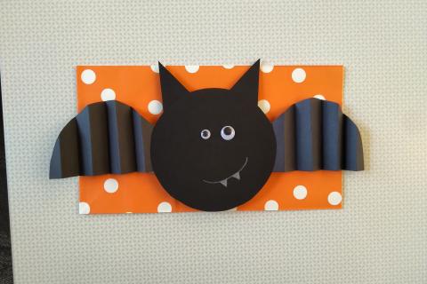 A black paper bat with googly eyes on an orange background