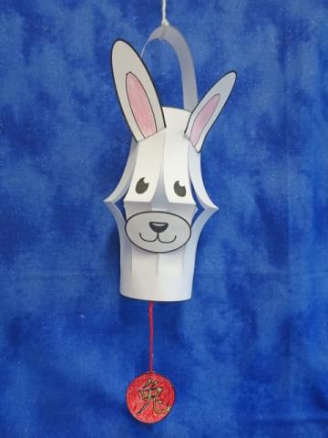 Paper lantern made with white paper with attached rabbit ears and face and red ornament hanging down from middle of lamp