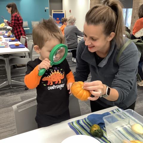 caregiver and child investigating a small pumpkin with a magnifying lense